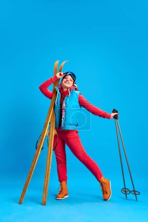 Foto de Chica joven en ropa de invierno de colores de pie con esquís sobre fondo de estudio azul. Vacaciones activas. Concepto de juventud, auto-expresión, vacaciones de invierno, estilo de vida activo, emociones - Imagen libre de derechos