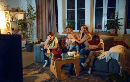 Family watching tv at home in the evening. Parents with children sitting on couch, watching horror movie, covering eyes in fear. Concept of family, leisure time, relaxation, childhood and parenthood