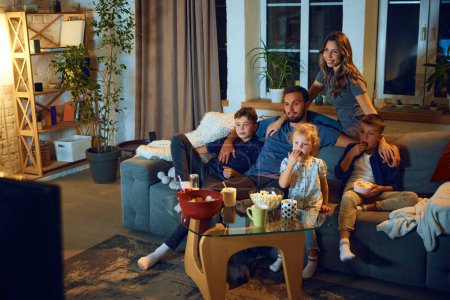 Photo for Happy loving family, parents and children sitting on couch in the evening at home and watching tv, eating snacks. Relaxation. Concept of family, leisure time, relaxation, childhood and parenthood - Royalty Free Image