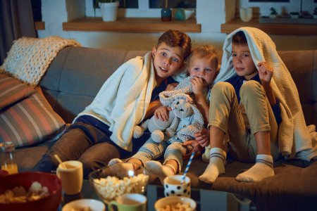 Photo for Emotional children, boys and little girl sitting on couch at home and watching tv, horror movie. Hiding under blanket. Concept of family, leisure time, relaxation, childhood and parenthood - Royalty Free Image