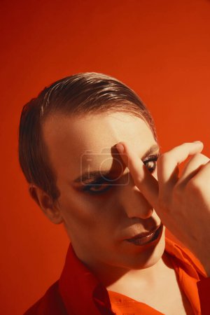 Téléchargez les photos : Portrait de jeune homme, transgenre, gay avec maquillage, en vêtements lumineux posant sur fond de studio rouge. Concept de maquillage masculin, mode, communauté lgbtq, identité de soi, acceptation - en image libre de droit