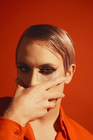 Foto de Queer. Retrato de un joven con maquillaje brillante y peinado posando sobre fondo rojo del estudio. Cosméticos. Concepto de maquillaje masculino, moda, comunidad lgbtq, identidad propia, aceptación - Imagen libre de derechos