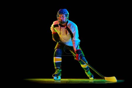 Foto de Chica joven, jugador de hockey en uniforme y entrenamiento casco, de pie con palo contra fondo negro estudio en luz de neón. Concepto de deporte profesional, competición, juego, acción, hobby - Imagen libre de derechos