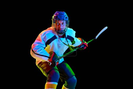 Photo for Concentrated, motivated young girl, hockey player standing in uniform with stick against black studio background in neon light. Concept of professional sport, competition, game, action, hobby - Royalty Free Image