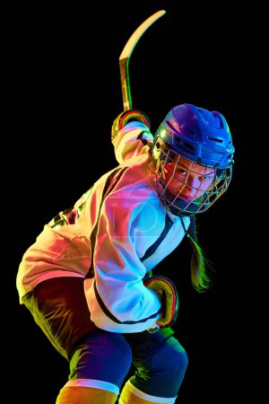 Foto de Chica joven concentrada y motivada, jugador de hockey de pie en uniforme con palo contra fondo de estudio negro en luz de neón. Concepto de deporte profesional, competición, juego, acción, hobby - Imagen libre de derechos