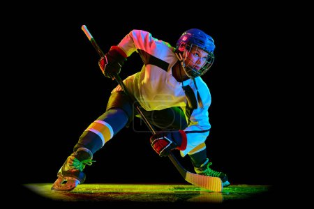 Photo for Concentrated young woman, hockey player un uniform and helmet, standing with stick against black studio background in neon light. Concept of professional sport, competition, game, action, hobby - Royalty Free Image