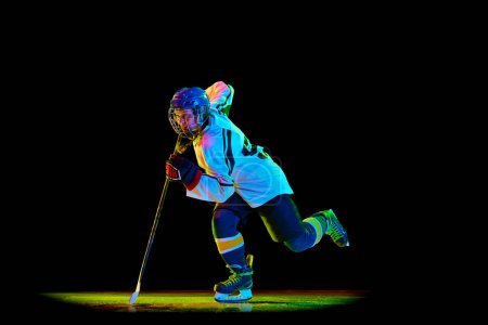 Téléchargez les photos : Image dynamique de jeune fille, joueur de hockey en uniforme et casque, en mouvement avec bâton sur fond de studio noir au néon. Concept de sport professionnel, compétition, jeu, action, passe-temps - en image libre de droit