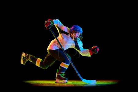 Foto de Jovencita competitiva, jugadora de hockey en movimiento, deslizándose sobre patines con arado, entrenando contra fondo de estudio negro en luz de neón. Concepto de deporte profesional, competición, juego, acción, hobby - Imagen libre de derechos