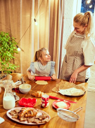 Photo for Happy family time. Young woman, mother cooking with little girl, daughter in modern kitchen at home. Cooking croissants and cookies. Concept of cookie day, motherhood, childhood, holidays, family - Royalty Free Image