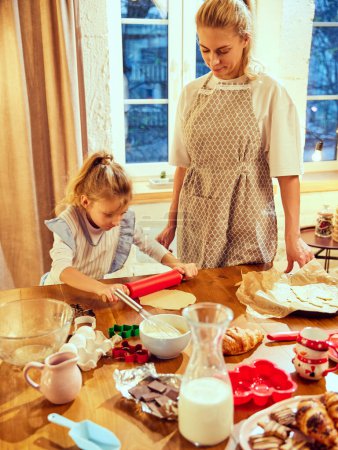 Foto de Mujer joven, madre de pie en la cocina por la noche con su hija pequeña, cocinar juntos, hacer masa, hornear croissants, galletas. Concepto de día de las galletas, maternidad, infancia, vacaciones, familia - Imagen libre de derechos