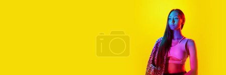 Téléchargez les photos : Portrait de jeune fille au visage sérieux, dans des vêtements modernes posant sur fond jaune avec réflexion néon. Concept d'art, style moderne, cyberpunk, futurisme et créativité. Bannière - en image libre de droit
