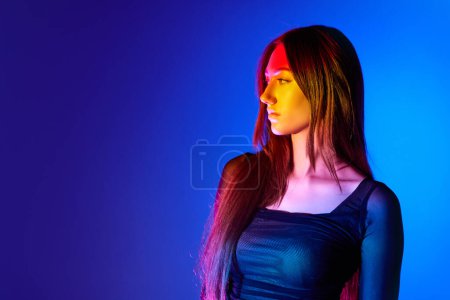 Foto de Retrato de una hermosa mujer joven en vestido elegante posando sobre fondo azul en luz de neón. Cara seria. Concepto de arte, estilo moderno, cyberpunk, futurismo y creatividad - Imagen libre de derechos