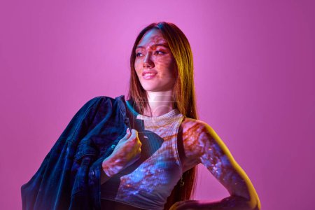 Foto de Retrato de mujer joven y elegante posando sobre fondo púrpura claro en luces de neón. Reflexión espacial. Concepto de arte, estilo moderno, cyberpunk, futurismo y creatividad - Imagen libre de derechos