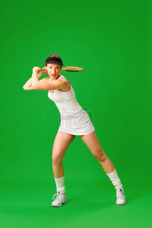 Téléchargez les photos : Jeune femme en tenue de sport blanche élégante jouant au tennis, s'entraînant sur fond de studio vert. Activité de week-end. Concept de sport, passe-temps, mode, jeunesse, émotions humaines, mode de vie - en image libre de droit
