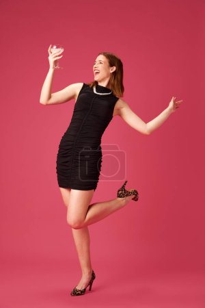 Téléchargez les photos : Joyeux jeune éléphant femme en robe noire célébrant, boire un cocktail et danser sur fond de studio rose. Concept de jeune, émotions humaines, style de vie, mode, fête, célébration - en image libre de droit