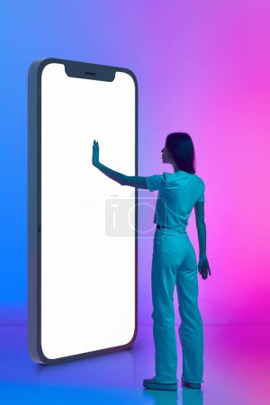 Foto de Mujer de pie cerca de gigante modelo 3D de teléfono, pantalla de desplazamiento contra degradado rosa azul estudio de fondo en luz de neón. Compras en línea. Concepto de emociones humanas, estilo de vida, cultura juvenil - Imagen libre de derechos