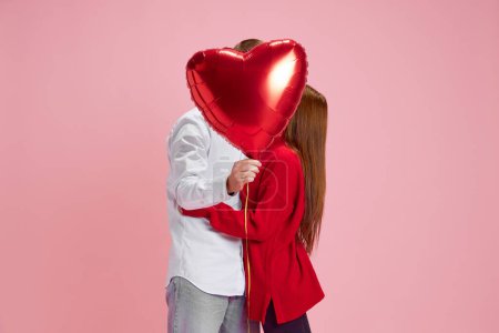 Téléchargez les photos : Joyeux jeune couple, femme et homme tenant un ballon en forme de cœur et s'embrassant derrière sur fond de studio rose. Concept d'amour, relation, Saint Valentin, émotions, style de vie - en image libre de droit