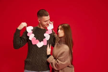 Photo for Celebrating holidays. Young happy loving couple, man and woman holding hearts, posing against red studio background. Concept of love, relationship, Valentines Day, emotions, lifestyle - Royalty Free Image
