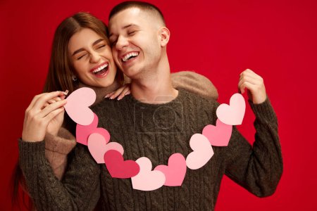 Photo for Happy, caring, loving young couple, boy and girl holding paper heart garland, celebrating holiday against red studio background. Concept of love, relationship, Valentines Day, emotions, lifestyle - Royalty Free Image