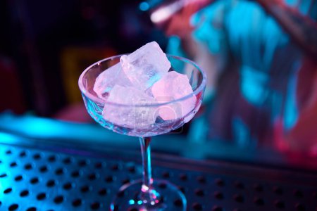 Photo for Focus on cocktail glass with ice cubes. Neon light. Modern bar. Preparing sweet and sour cocktails. Concept of occupation, nightlife, bar, party, alcohol drink, mixologist - Royalty Free Image
