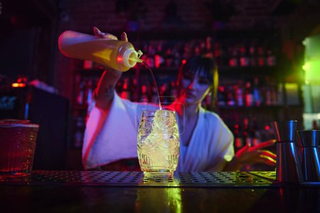 Foto de Hermosa joven que trabaja como camarera en un bar moderno, haciendo un delicioso cóctel, agregando líquidos en el vaso con hielo. Concepto de ocupación, vida nocturna, bar, fiesta, bebida alcohólica, mixólogo - Imagen libre de derechos