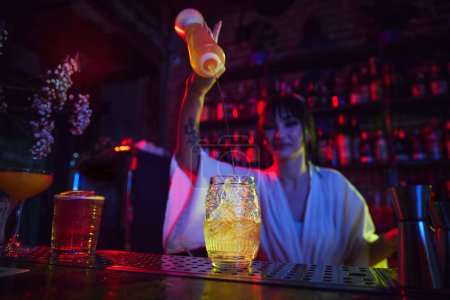 Photo for Beautiful young girl working as bartender in modern bar, making delicious cocktail, adding liquids into glass with ice. Concept of occupation, nightlife, bar, party, alcohol drink, mixologist - Royalty Free Image