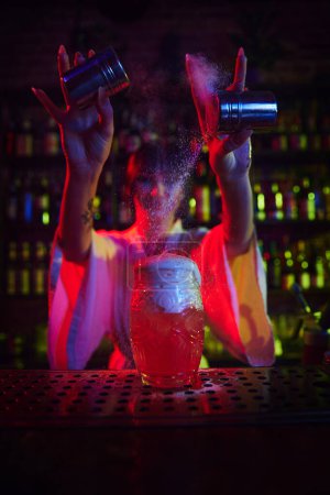 Foto de Hermosa joven que trabaja como camarera en el bar moderno, haciendo delicioso cóctel, añadiendo polvo de canela en espuma. Concepto de ocupación, vida nocturna, bar, fiesta, bebida alcohólica, mixólogo - Imagen libre de derechos