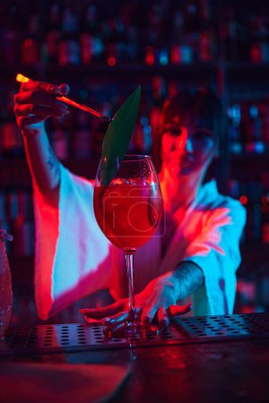 Foto de Joven mujer hermosa, barman en bar moderno, haciendo delicioso cóctel Aperol Spritz en neón, decorando con hojas. Concepto de ocupación, vida nocturna, bar, fiesta, bebida alcohólica, mixólogo - Imagen libre de derechos