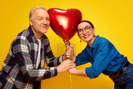 Photo for Beautiful, happy, senior couple, man and woman celebrating romantic holiday, holding balloon in heart shape over yellow background. Concept of marriage, relationship, Valentines Day, love, emotions - Royalty Free Image