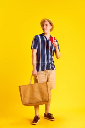 Téléchargez les photos : Jeune homme élégant et souriant en chemise et short, tenant son sac, buvant de la coke sur fond de studio jaune. Touristique. Concept d'émotions humaines, jeunesse, vacances d'été, mode, temps libre - en image libre de droit