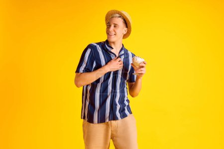 Foto de Guapo joven sonriente en camisa a rayas, pantalones cortos y sombrero de paja comiendo delicioso helado de vainilla sobre fondo de estudio amarillo. Concepto de emociones humanas, juventud, vacaciones de verano, moda - Imagen libre de derechos
