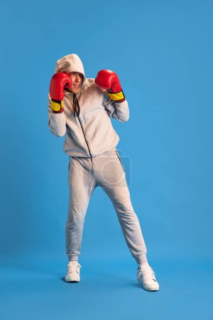 Photo for Young guy in grey sportswear standing in red boxing gloves, training against blue studio background. Fighter. Concept of human emotions, youth, sportive lifestyle, fashion, combat sport - Royalty Free Image