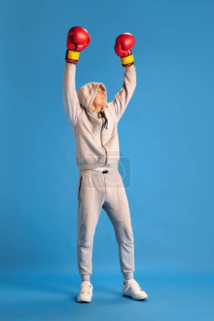 Téléchargez les photos : Jeune homme en vêtements de sport gris debout dans des gants de boxe rouges avec les mains levées et sourire sur fond bleu studio. Champion. Concept d'émotions humaines, de jeunesse, de style de vie sportif, de mode - en image libre de droit