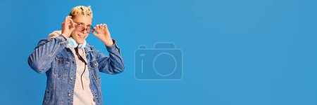 Téléchargez les photos : Portrait de beau jeune homme en lunettes de soleil roses posant sur fond bleu studio. Concept d'émotions humaines, de jeunesse, de positivité, de mode décontractée, de musique. Bannière. Espace vide pour insérer du texte - en image libre de droit
