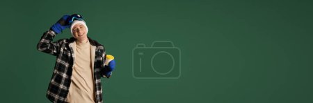 Photo for Young smiling guy in checkered jacket, hat, goggles standing with coffee on green background. Concept of human emotions, youth, winter fashion, vacations holidays. Banner. Empty space to insert text - Royalty Free Image