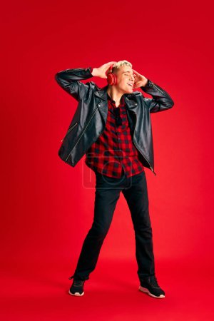 Foto de Elegante joven con camisa a cuadros y chaqueta de cuero escuchando música en auriculares y bailando sobre fondo rojo del estudio. Concepto de emociones humanas, juventud, moda, estilo de vida, positividad - Imagen libre de derechos