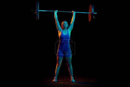 Téléchargez les photos : Image complète de l'athlète professionnel, entraînement de la jeune femme, levage de poids lourd, haltère sur fond noir dans la lumière au néon. Concept de sport, force, mode de vie sain, puissance, endurance - en image libre de droit