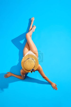 Téléchargez les photos : Vue de dessus. Élégante jeune femme au corps mince, en forme, bronzé, portant un maillot de bain et un chapeau de paille, couchée sur fond de studio bleu. Concept d'été, vacances, repos, vacances, mode et beauté - en image libre de droit