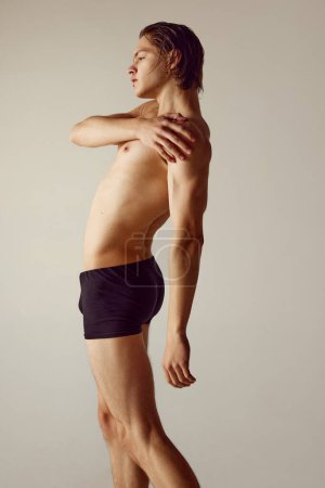 Photo for Side view. Handsome shirtless young guy with muscular body standing in underwear against grey studio background. Fitness. Concept of mens beauty, health, body care, sportive lifestyle - Royalty Free Image