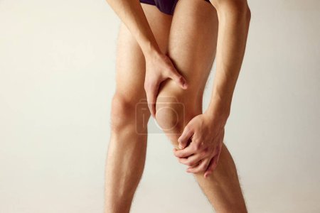 Téléchargez les photos : Image recadrée de jambes masculines sur fond de studio gris. Homme tenant le genou, souffrant de douleur. Inflammation. Concept d'hommes beauté, santé, soins du corps, style de vie sportif - en image libre de droit