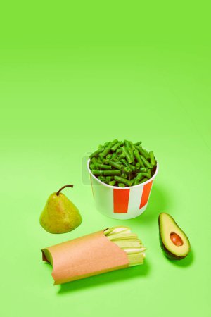 Photo for Healthy snacks. Green beans into popcorn basket, avocado, celery and pear against green background. Concept of healthy food, nutrition, diet, organic products, diet, vegetable vitamins. Poster, ad - Royalty Free Image