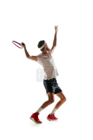 Téléchargez les photos : Pleine longueur de jeune homme en vêtements de sport, joueur de tennis pratiquant, frappant balle avec raquette isolé sur fond blanc. Concept de sport, passe-temps, mode de vie actif et sain, compétition - en image libre de droit