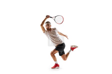 Téléchargez les photos : Image dynamique d'un jeune homme concentré, joueur de tennis pratiquant, jouant, frappant une balle avec raquette isolée sur fond blanc. Concept de sport, passe-temps, mode de vie actif et sain, compétition - en image libre de droit