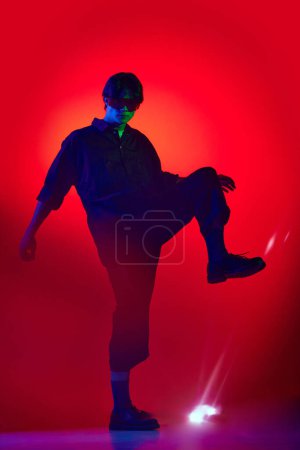 Photo for Full-length of handsome, extraordinary young guy in jumpsuit posing over red background in neon light. Fashion show. Concept of youth culture, fashion and male beauty, emotions, inspiration, trends - Royalty Free Image