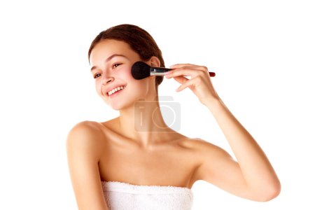 Photo for Natural makeup look. Young smiling beautiful girl with smooth, well-kept, spotless skin applying blush on cheeks over white background. Concept of natural beauty, skin care, cosmetology and cosmetics - Royalty Free Image