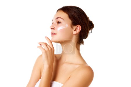 Photo for Side view image of young beautiful girl with bare shoulders standing with moisturizing cream on face against white background. Concept of natural female beauty, skin care, cosmetology and cosmetics - Royalty Free Image
