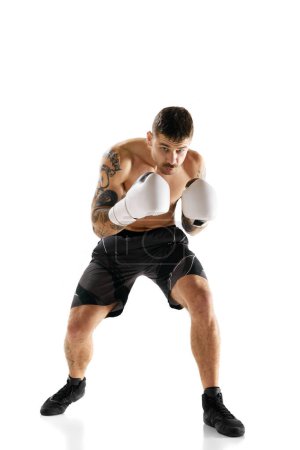 Téléchargez les photos : Image pleine longueur du jeune homme musclé, athlète de boxe en mouvement, s'entraînant isolé sur fond blanc. Concept de sport professionnel, sport de combat, arts martiaux, force - en image libre de droit
