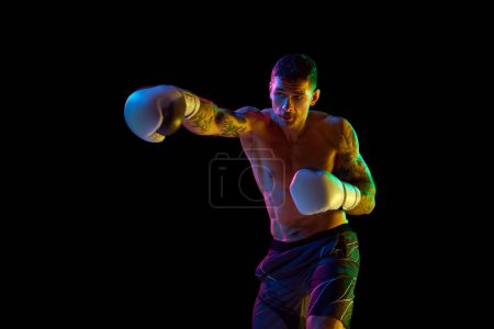 Téléchargez les photos : Jeune homme avec torse nu, fort, musclé entraînement du corps, boxe athlète poinçonnage isolé sur fond noir dans la lumière au néon. Concept de sport professionnel, sport de combat, arts martiaux, force - en image libre de droit