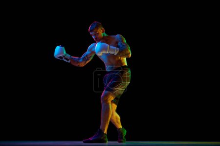 Téléchargez les photos : Image pleine longueur du jeune homme athlétique, boxeur musclé et torse nu en mouvement, se battant, pratiquant des crochets isolés sur fond noir au néon. Concept de sport, sport de combat, arts martiaux, force - en image libre de droit
