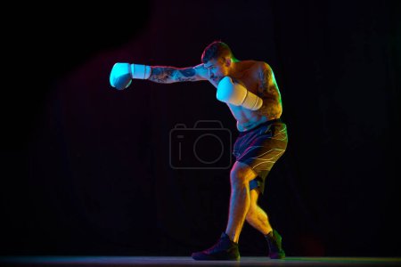 Téléchargez les photos : Jeune homme fort et musclé, athlète de boxe en gants, s'entraînant, se battant isolé sur fond noir sous la lumière du néon. Concept de sport professionnel, sport de combat, arts martiaux, force - en image libre de droit
